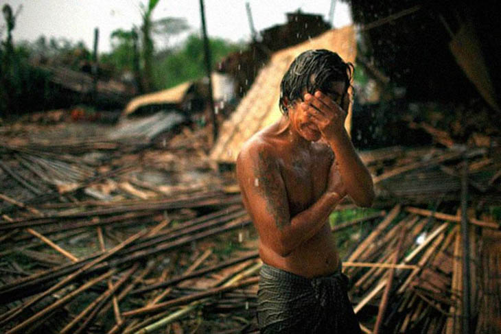 Hhaing The Yu, 29, holds his face in his hand as rain falls on the decimated remains of his home